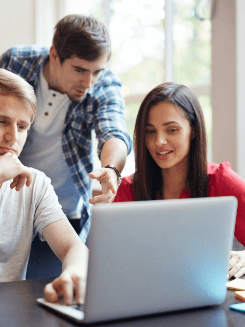 Three young professionals—two men and one woman—gathered around a silver laptop in a bright workspace. One man points at the screen while the woman in a red shirt speaks, and colorful sticky notes and markers sit on the table nearby.