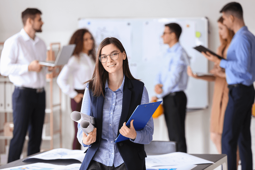 A young woman with glasses and long brown hair smiles at the camera while holding a blue clipboard and rolled-up architectural blueprints. In the blurred background, colleagues collaborate in a modern, bright office space with whiteboards.