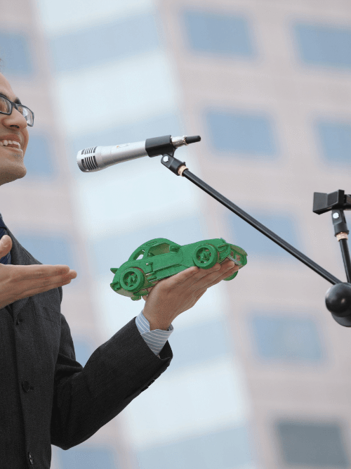A confident student at CATS Pakistan giving a public speech and presenting a green 3D car model during a technical innovation seminar.