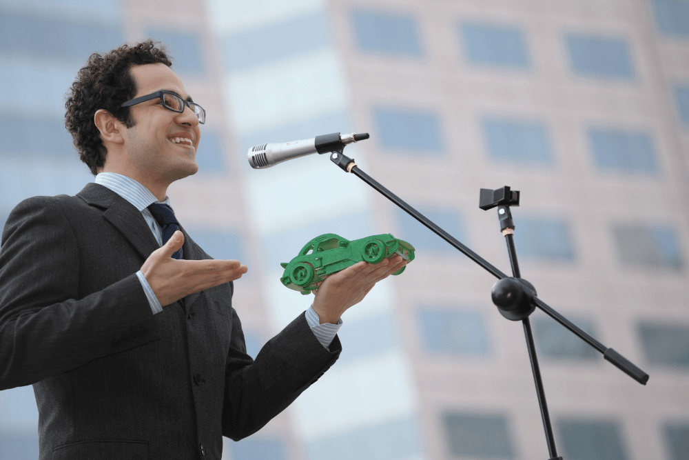 A confident student at CATS Pakistan giving a public speech and presenting a green 3D car model during a technical innovation seminar.